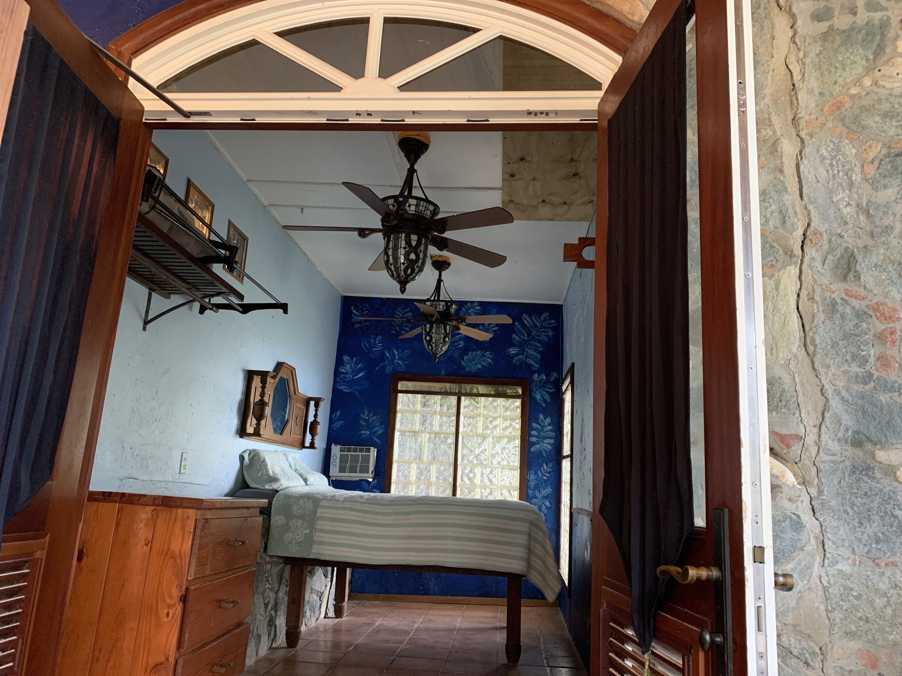 A bedroom with a bed, wooden dresser, ceiling fan, and a window with decorative glass.