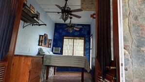 Quarto com cama, cómoda de madeira, ventoinha no teto e janela com vidro decorativo.