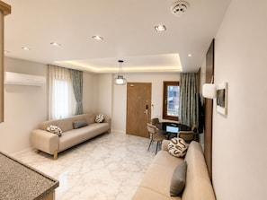 A modern living room with a sofa, dining table, and chairs. There is a wooden door and a window with curtains. - Auga Apart (Çaykara)