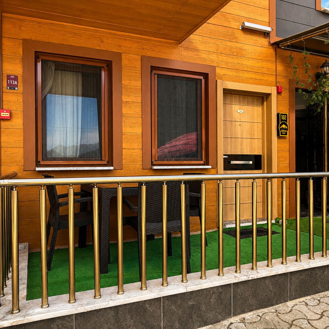 A wooden building with two windows, a door, and a small outdoor area with artificial grass and a metal railing.