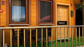 A wooden building with two windows, a door, and a small outdoor area with artificial grass and a metal railing.
