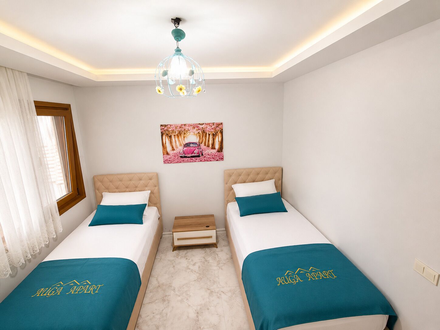 A hotel room with two beds, a chandelier, a picture on the wall, and a window with curtains.