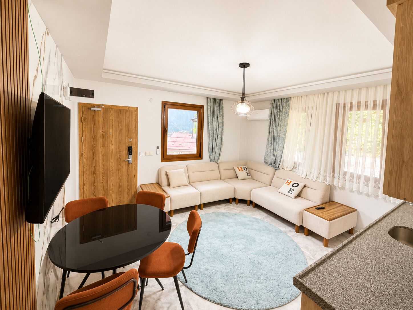 A modern living room with a sectional sofa, a dining table with chairs, a TV mounted on the wall, and a kitchen area with a sink.