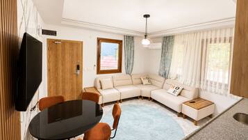 A modern living room with a sectional sofa, a dining table with chairs, a TV mounted on the wall, and a kitchen area with a sink.