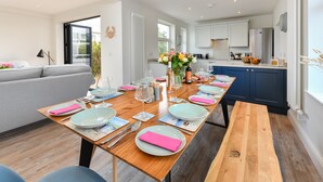 A modern dining area with a wooden table set for four, surrounded by a grey sofa and a kitchen in the background. - Beautifully refurbished holiday home in the centre of Abersoch with parking. (Abersoch)