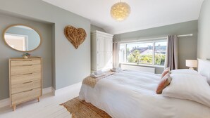 A bedroom with a bed, a dresser, a mirror, a heart-shaped decoration, and a window with a view of houses. - Beautifully refurbished holiday home in the centre of Abersoch with parking. (Abersoch)