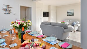 A dining area with a wooden table set for a meal, a grey sofa in the background, and a TV mounted on the wall. - Beautifully refurbished holiday home in the centre of Abersoch with parking. (Abersoch)