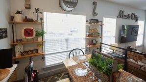 Un coin repas avec une table dressée pour deux personnes, entouré d’étagères remplies de divers objets et d’une grande fenêtre équipée de stores.