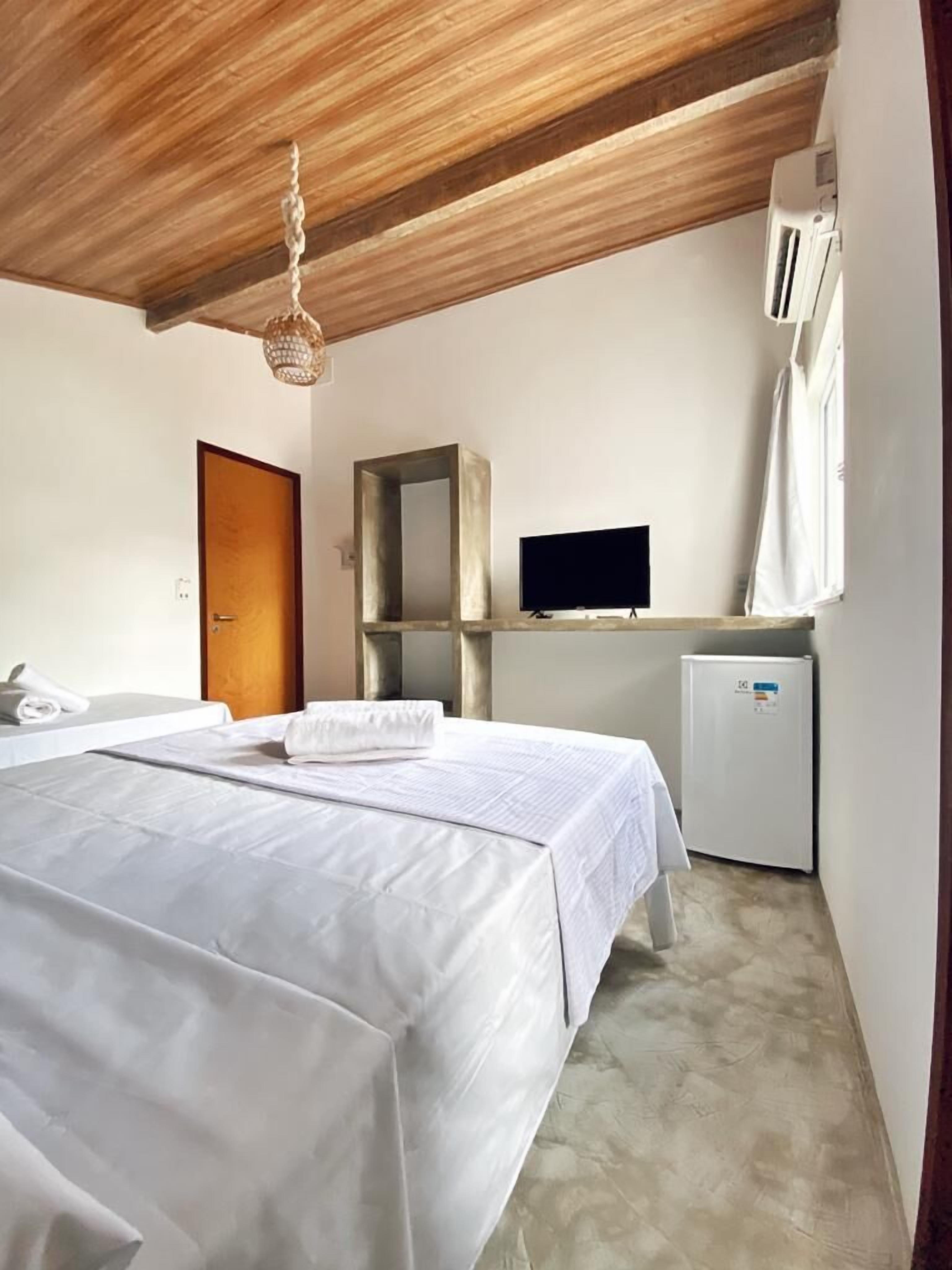 A hotel room with a wooden ceiling, a bed with white linens, a television, a small refrigerator, and a window with curtains.