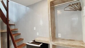 Una habitación con una escalera de madera, un cubo de basura negro y una estantería de madera con perchas para ropa.