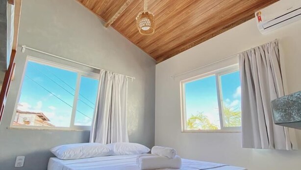 Un dormitorio con techo de madera, una cama con ropa de cama blanca, dos ventanas con cortinas y un equipo de aire acondicionado.