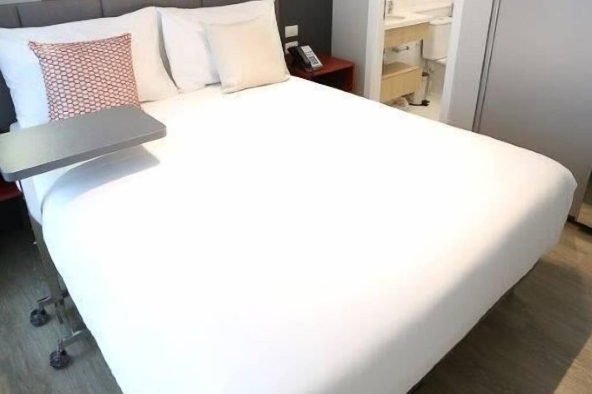 A hotel room with a large bed, a bedside table, a desk, an air conditioning unit, and two framed pictures on the wall.