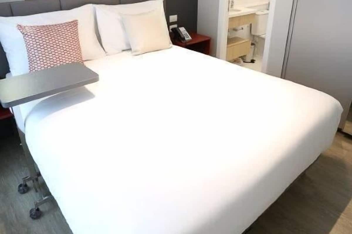 A hotel room with a large bed, a bedside table, a desk, and a chair. There are two framed pictures on the wall, an air conditioning unit, and a door leading to a bathroom.