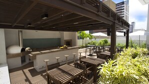 An outdoor patio area with wooden tables and chairs, a bar with a countertop and stools, and potted plants. - Astron Suítes Mondial Salvador by Nobile (Salvador)