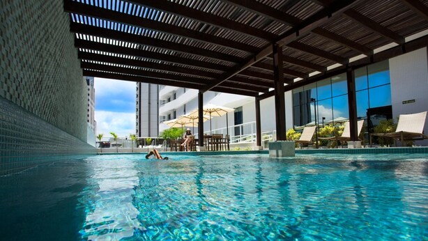 An indoor swimming pool with clear water, a covered roof, and a view of a building with large windows and outdoor umbrellas. - Astron Suítes Mondial Salvador by Nobile (Salvador)