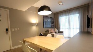 A modern hotel room with a wooden dining table, white chairs, a black pendant light, a sofa with cushions, a large window with curtains, and a flat-screen TV mounted on the wall. - Astron Suítes Mondial Salvador by Nobile (Salvador)