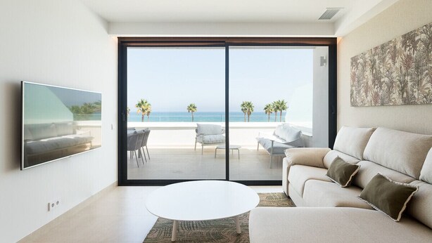 Una sala de estar moderna con un amplio ventanal corredero que da a un balcón con vistas al mar y palmeras.