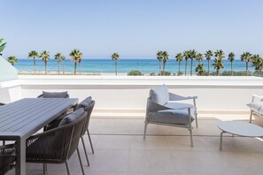 An outdoor terrace with a table and chairs, a small round table, and a view of the beach with palm trees. - ApartUP Denia Beachfront Deluxe (Denia)