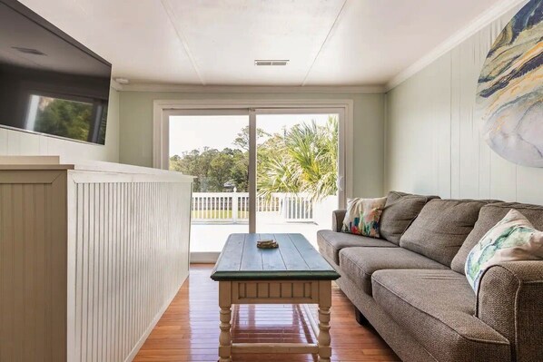 A living room with a wooden table, a sofa, and a view of the outdoors. - The Cosmic Folly Space Ranch (Folly Beach)