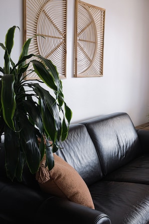 Um sofá de couro preto com uma almofada marrom, uma grande planta verde e duas obras de arte abstratas penduradas na parede.