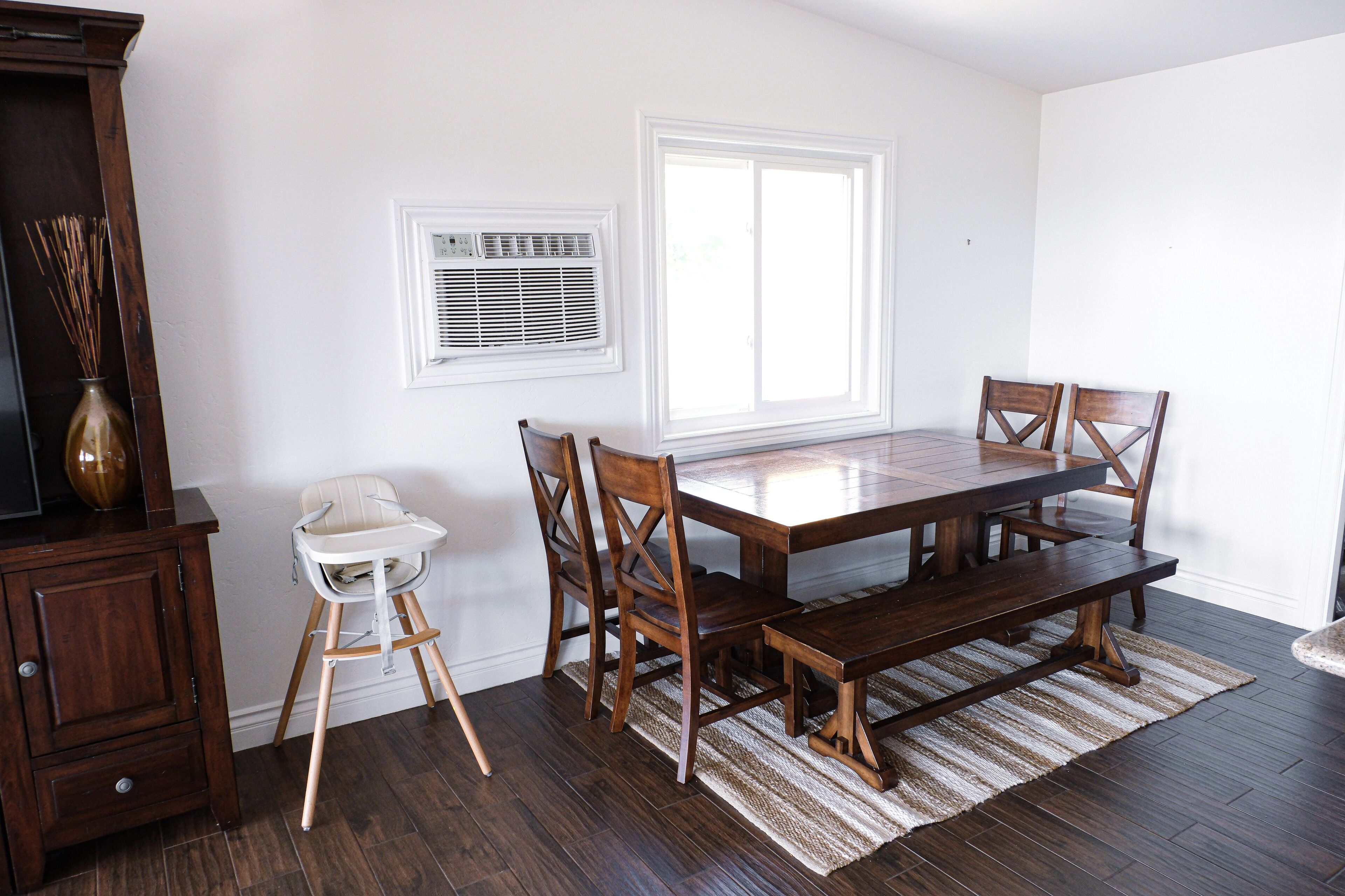 A dining area with a wooden table and chairs, a white high chair, a wooden cabinet, and a large window with blinds.