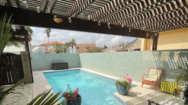 A pool area with a tiled floor, a pool, and a pergola overhead. There are chairs and potted plants around the pool. - North Padre Island waterfront canal  home w/private dock and pool (Corpus Christi)