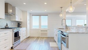 Une cuisine moderne avec des meubles blancs, une cuisinière à gaz, un four encastré, un lave-vaisselle et une grande fenêtre donnant sur la mer.