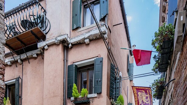 A narrow street with buildings featuring balconies with plants, a red banner, and a flag. - Lo Squero 2 Charming Apartment in Dorsoduro (Venice)