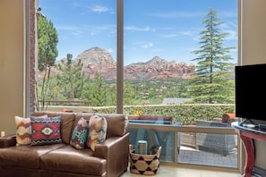 A living room with a leather couch, patterned cushions, a flat-screen TV, and a large window overlooking a mountainous landscape.