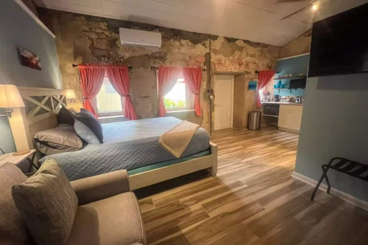 A bedroom with a bed, a sofa, a stone wall, and a kitchen area.