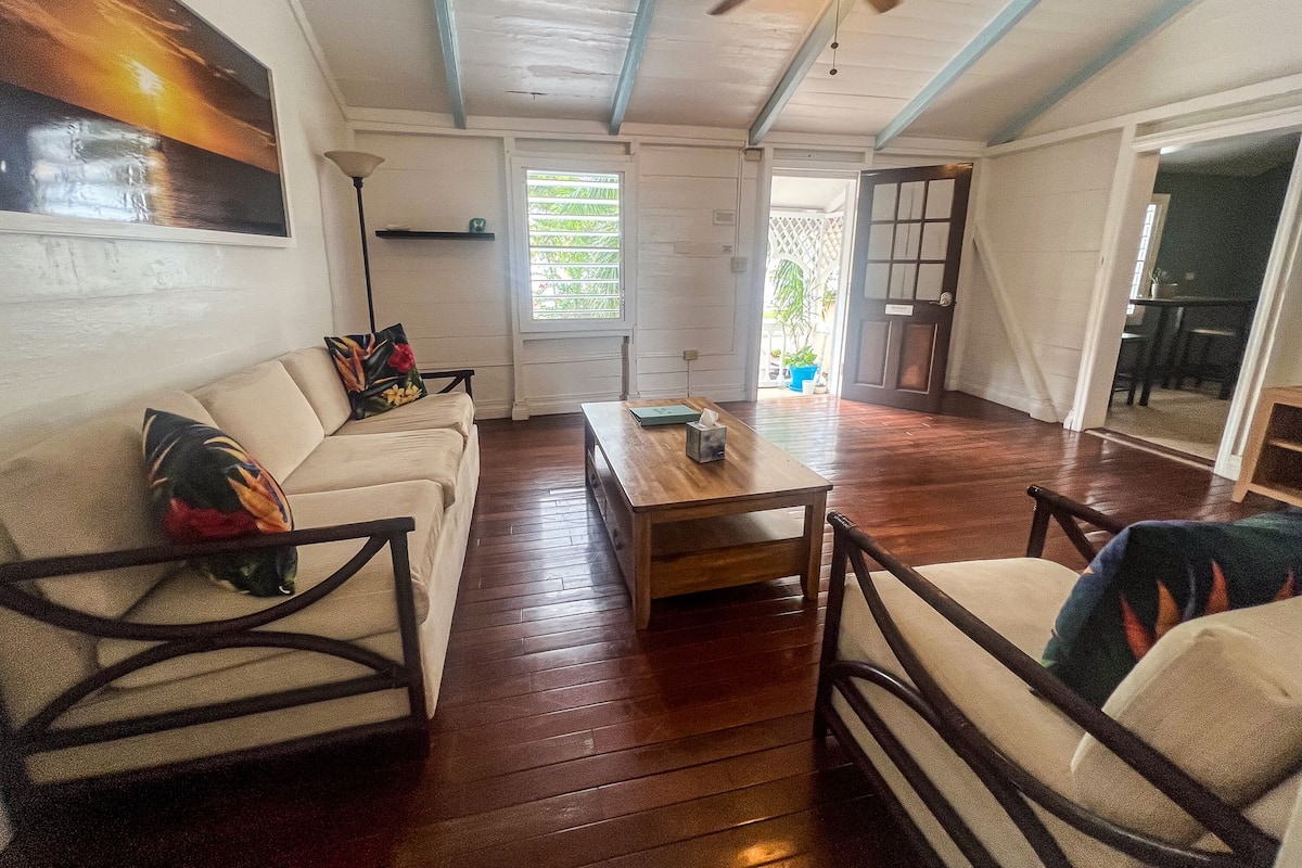 A cozy living room with a sofa, coffee table, and chairs. There is a ceiling fan, a window with blinds, and a door leading to another room.