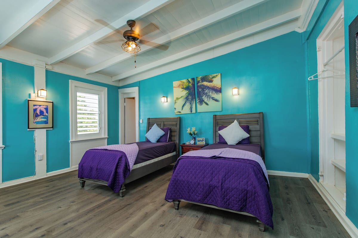 A room with two beds, teal walls, wooden flooring, ceiling fan, and wall-mounted lights.