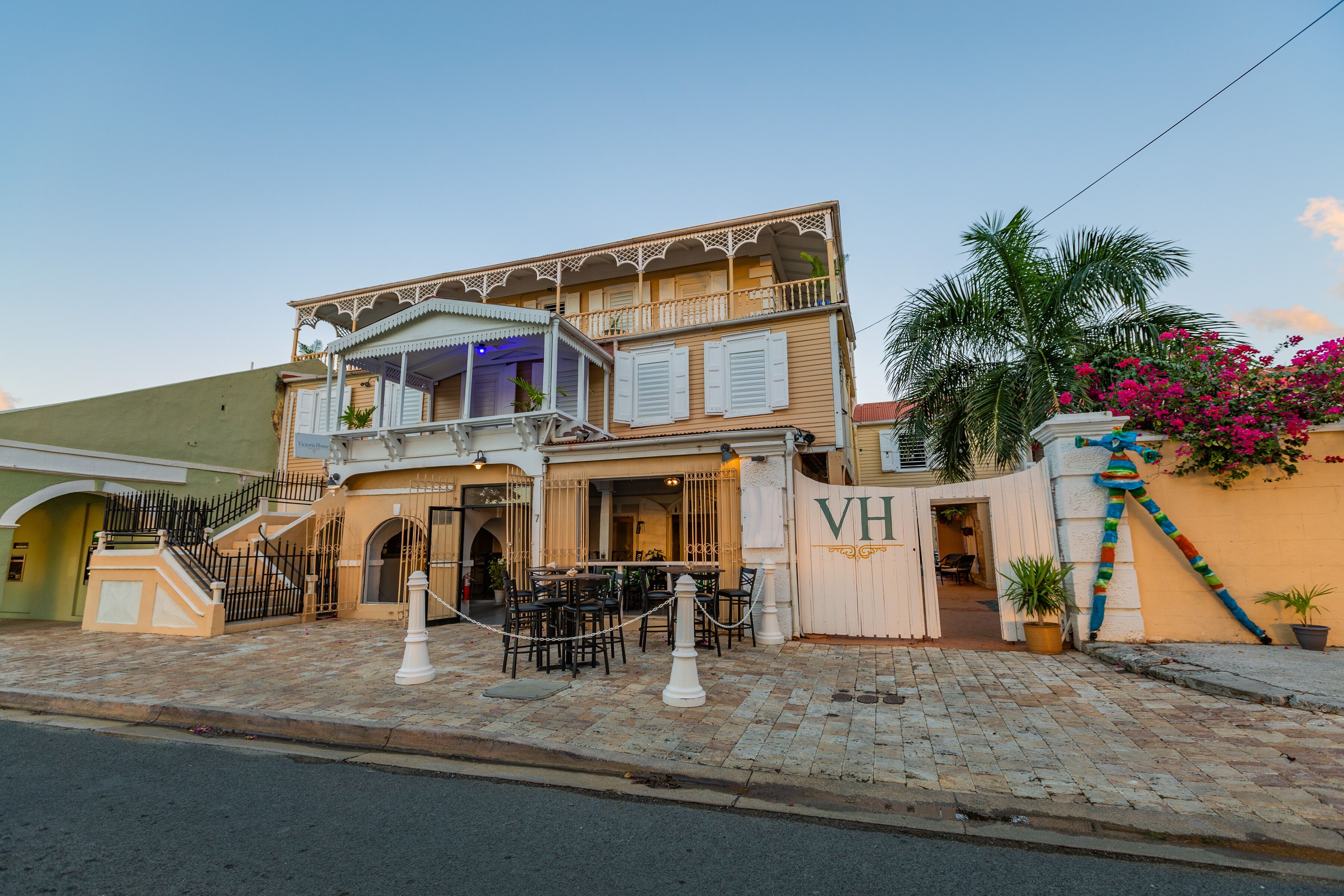A two-story building with a balcony, outdoor seating area, and a gate with the letters "VH".