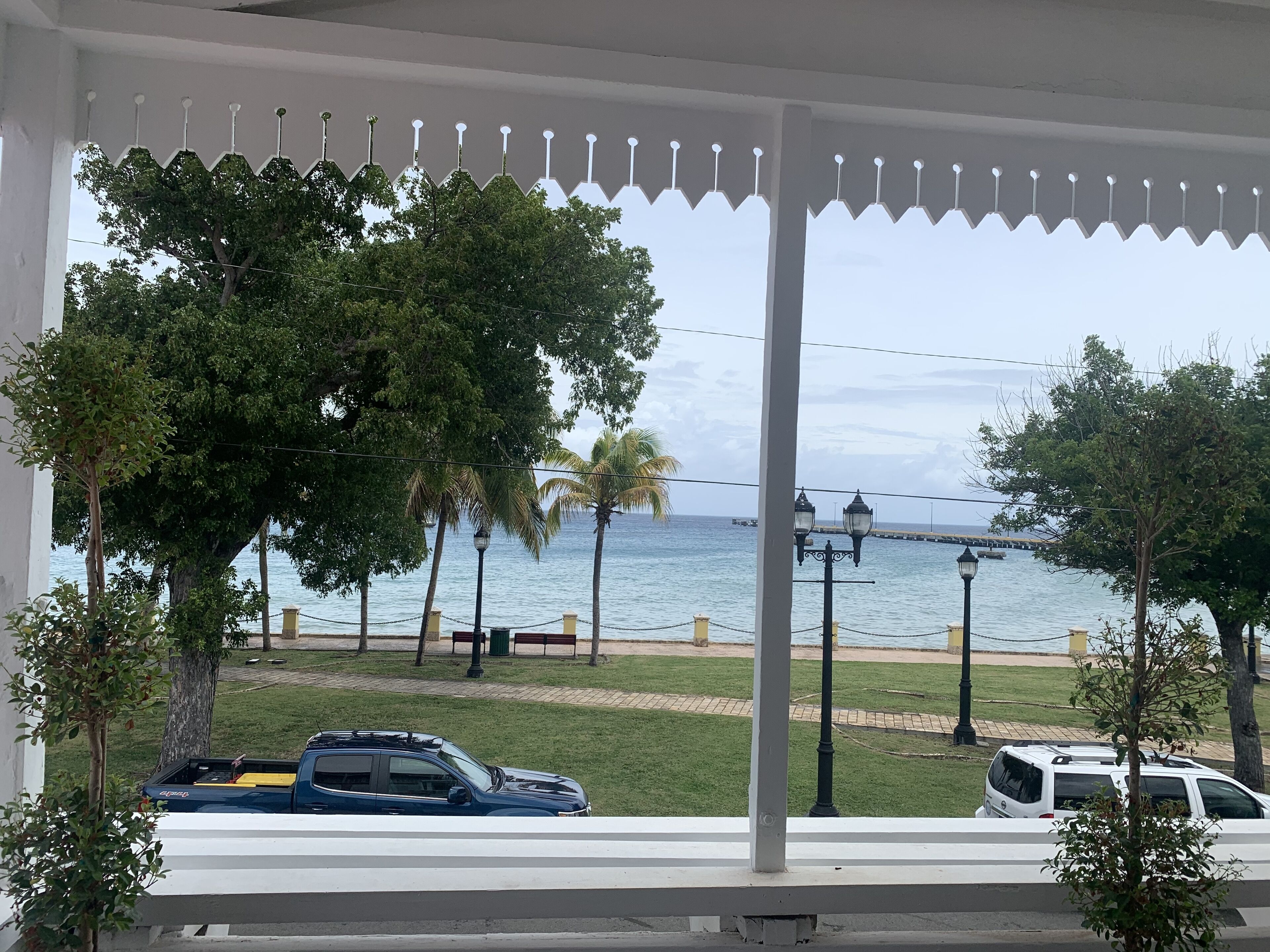 A covered outdoor area with a view of the sea, featuring a white railing, trees, and parked cars.