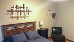 Un dormitorio con cabecera de madera, cama con ropa de cama azul, mesita de noche con lámpara, sillón de cuero negro y dos cuadros en la pared.