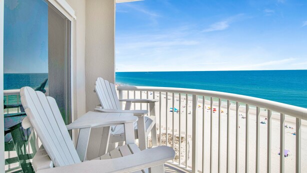 Ein Balkon mit weißen Stühlen und einem Tisch mit Blick auf einen Strand, auf dem sich Personen und Sonnenschirme befinden.