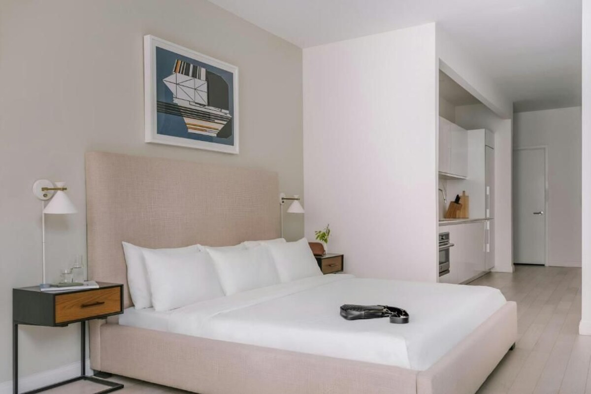 A modern hotel room with a large bed, bedside tables, a nightstand, a framed picture on the wall, and a view into the adjacent room.