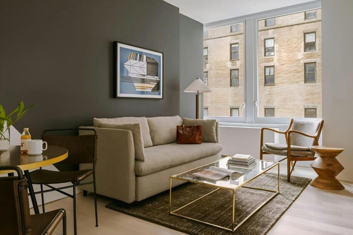 A modern living room with a sofa, coffee table, and chairs, a painting on the wall, and a view of a brick building through a window.