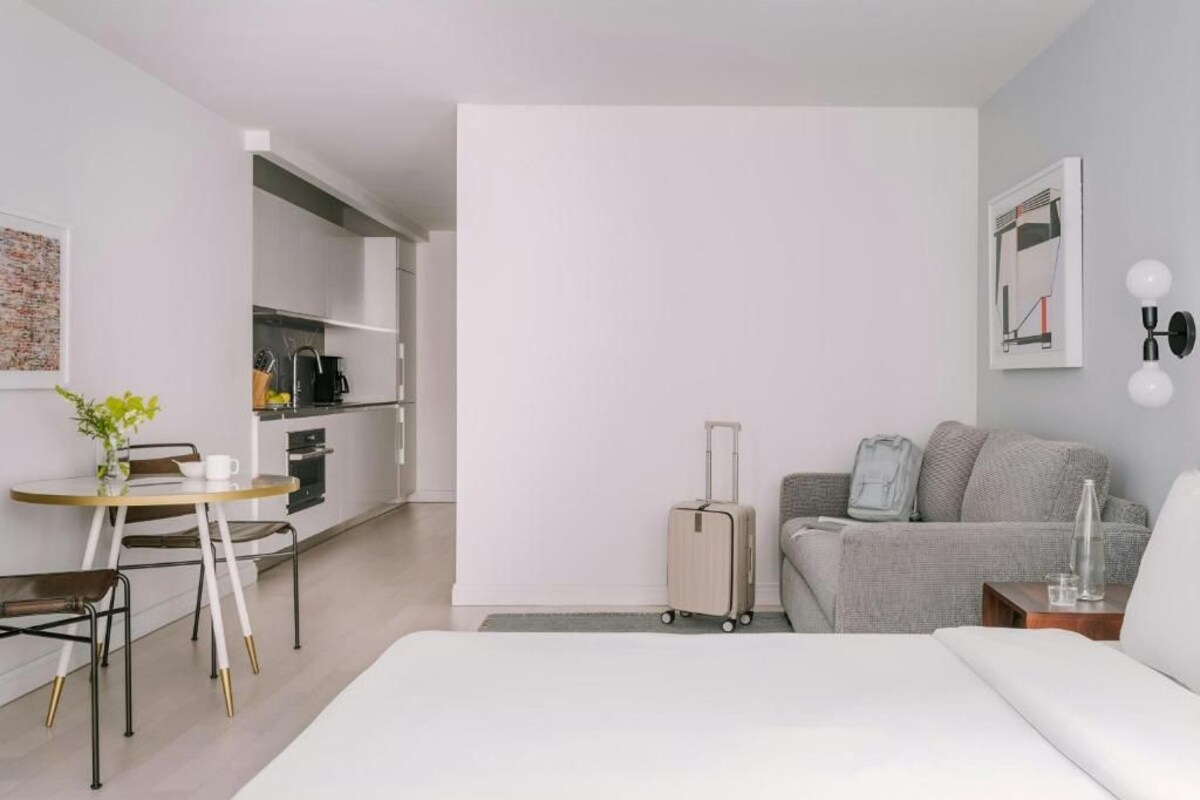 A modern hotel room with a bed, a small dining table with chairs, a kitchenette, and a grey sofa.