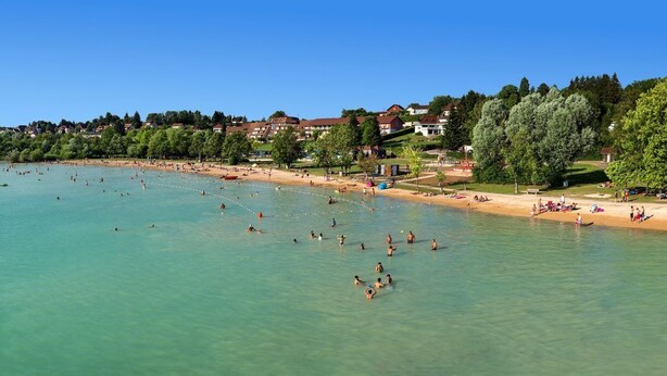 Een strand met helder water, mensen die zwemmen en zonnebaden, en huizen op de achtergrond.