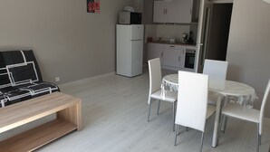 Een ruimtelijk ingerichte leefruimte met een bank, een eettafel met stoelen, een keukenhoek met kasten en apparatuur, en een kleine koelkast.