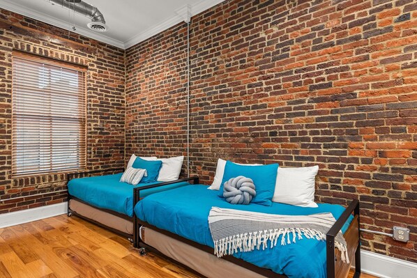 A room with a brick wall, a bed with blue bedding, and a window with blinds.