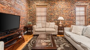 A living room with a brick wall, a flat-screen TV, a beige sofa, a glass coffee table, and a lamp. - The Johnny Cash Dual Condos Downtown at Printer's Alley & Steps to Broadway (Nashville)