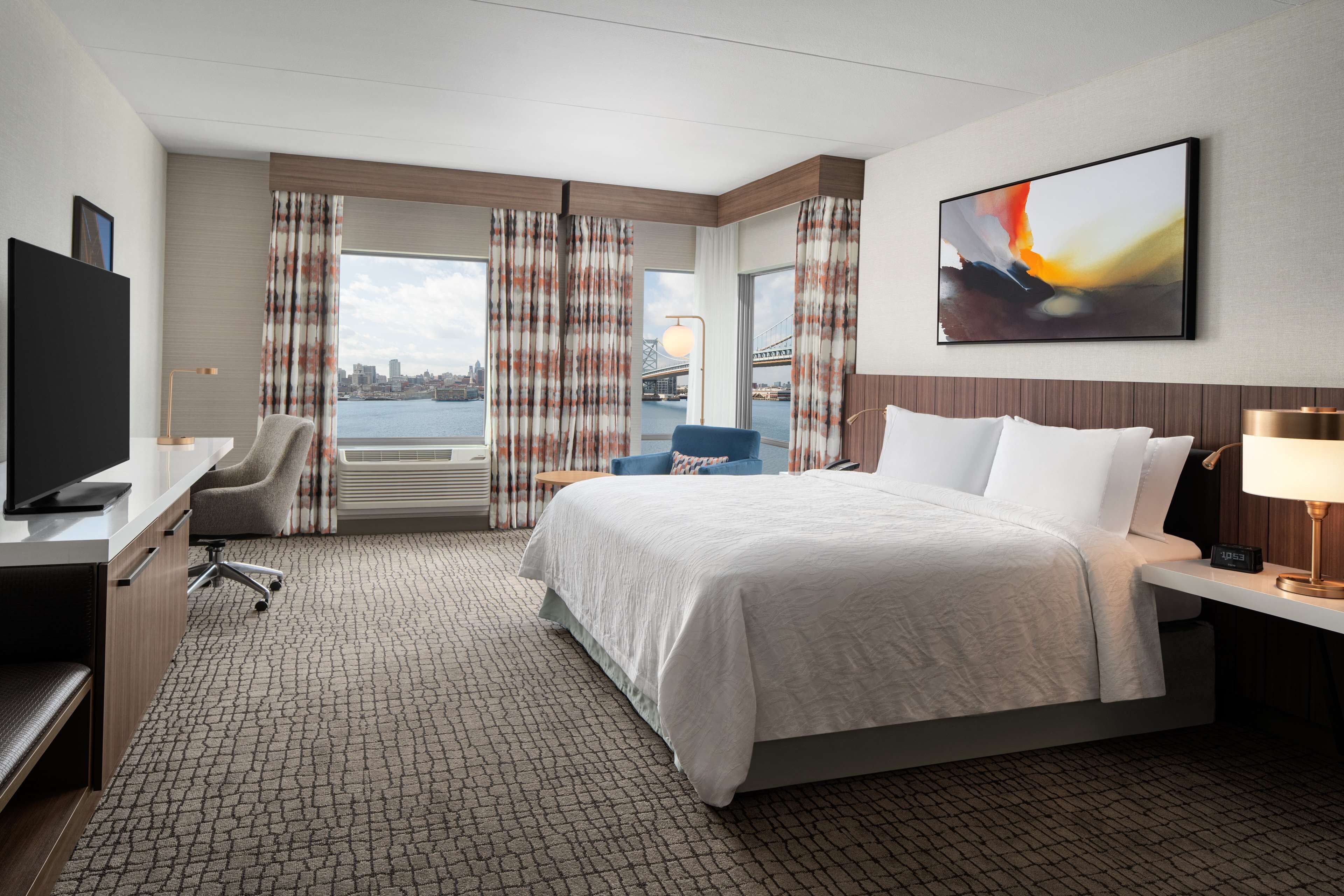 A hotel room with a bed, a desk with a chair, a TV, and a view of a bridge and cityscape.