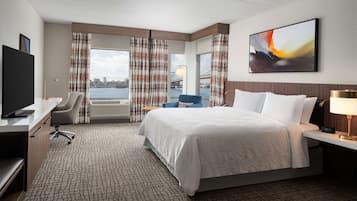 A hotel room with a bed, a desk with a chair, a TV, and a view of a bridge and cityscape.