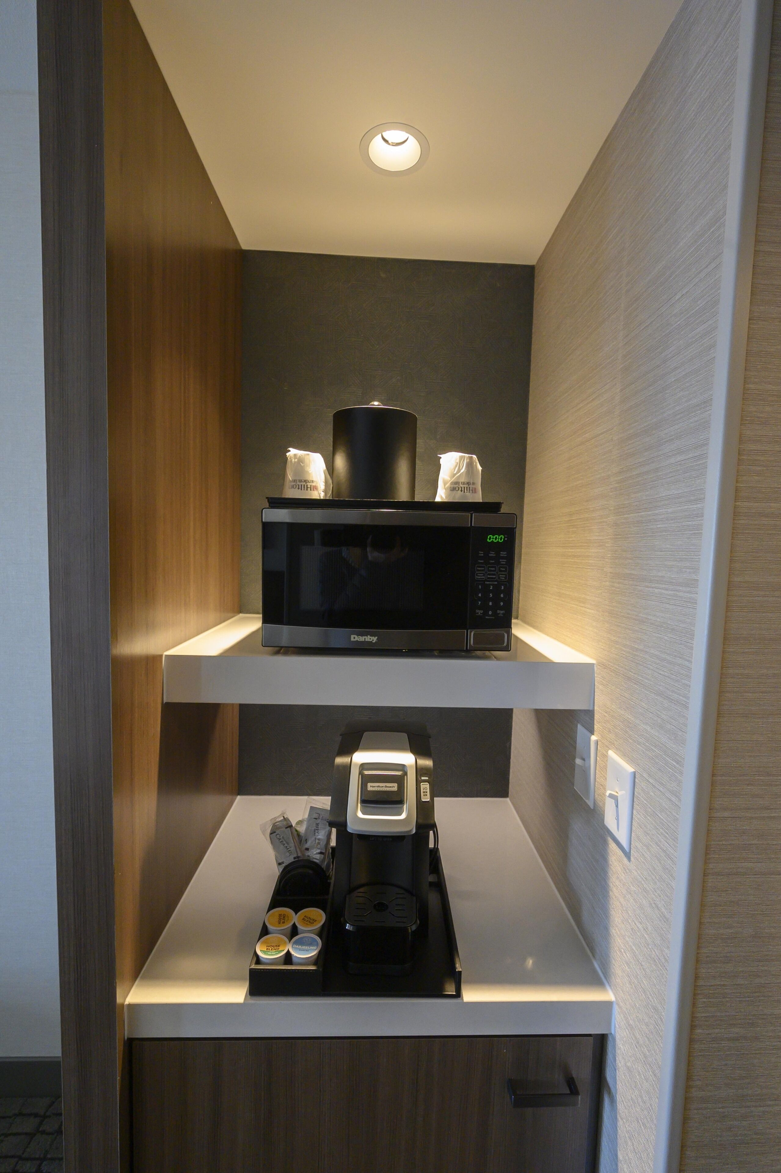 A compact kitchenette with a microwave and coffee maker on a white countertop, a small storage drawer below, and a wooden panel on the left.