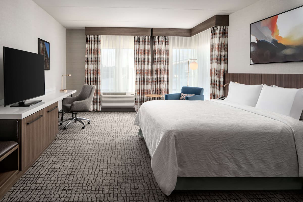 A hotel room with a large bed, a desk with a flat-screen TV, a chair, and a window with curtains.
