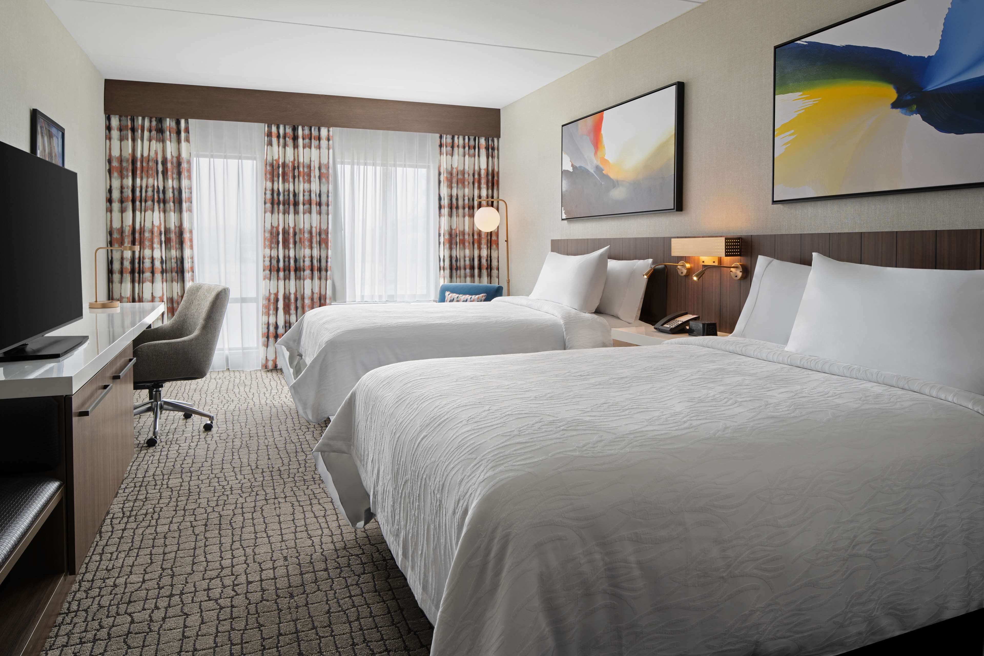 A hotel room with two beds, a desk with a chair, a TV, and a large window with curtains.