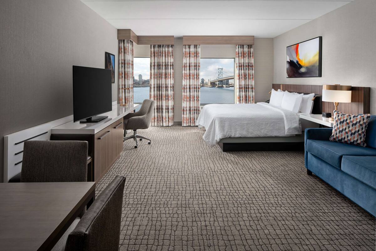 A hotel room with a bed, a desk with a TV, a chair, a sofa, and a view of a bridge and water.
