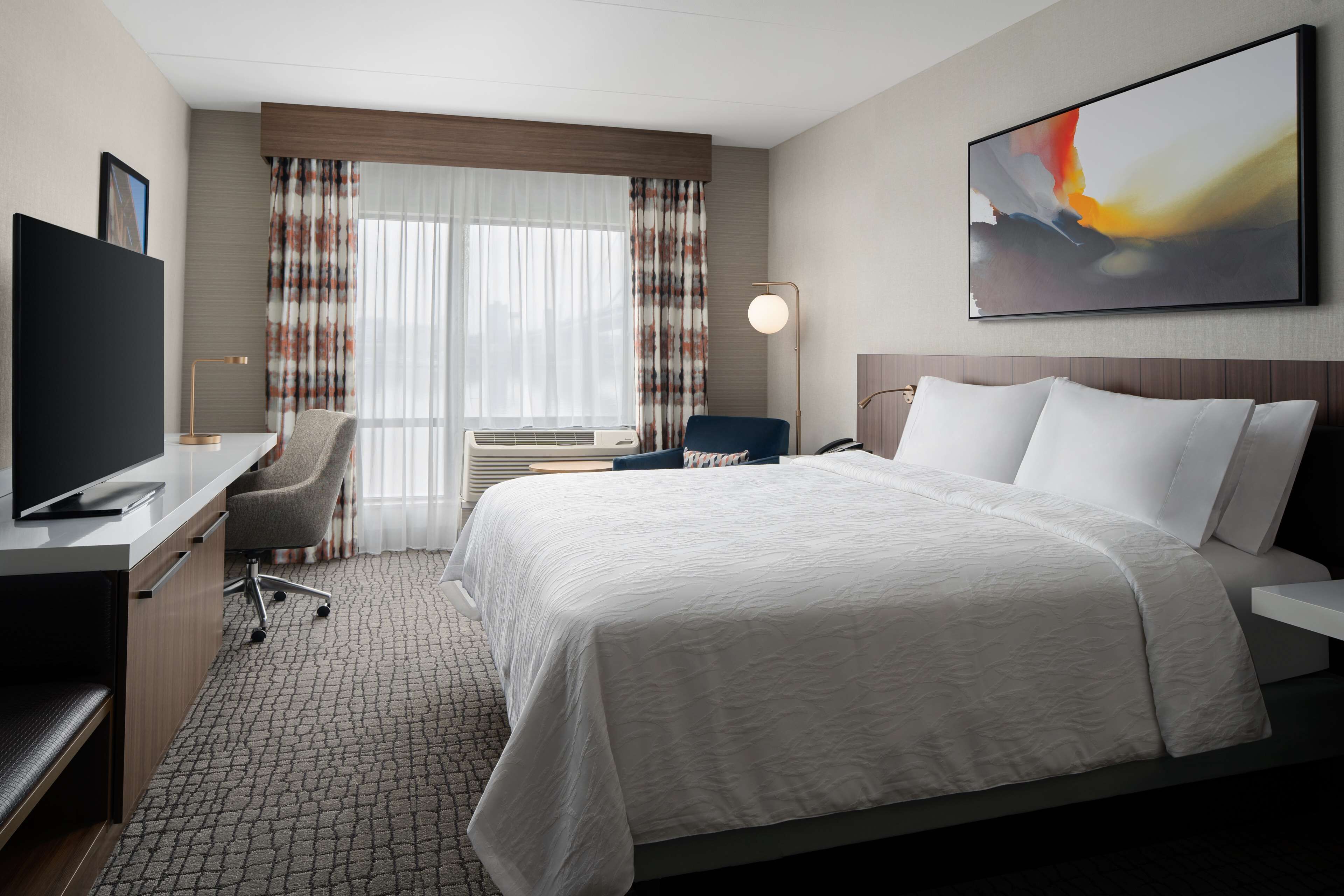 A hotel room with a bed, a desk with a TV, a chair, a lamp, and a painting on the wall.